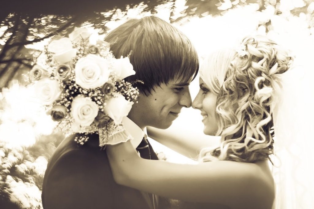 Bride and groom smiling and embracing under trees on their wedding day with bouquet in view