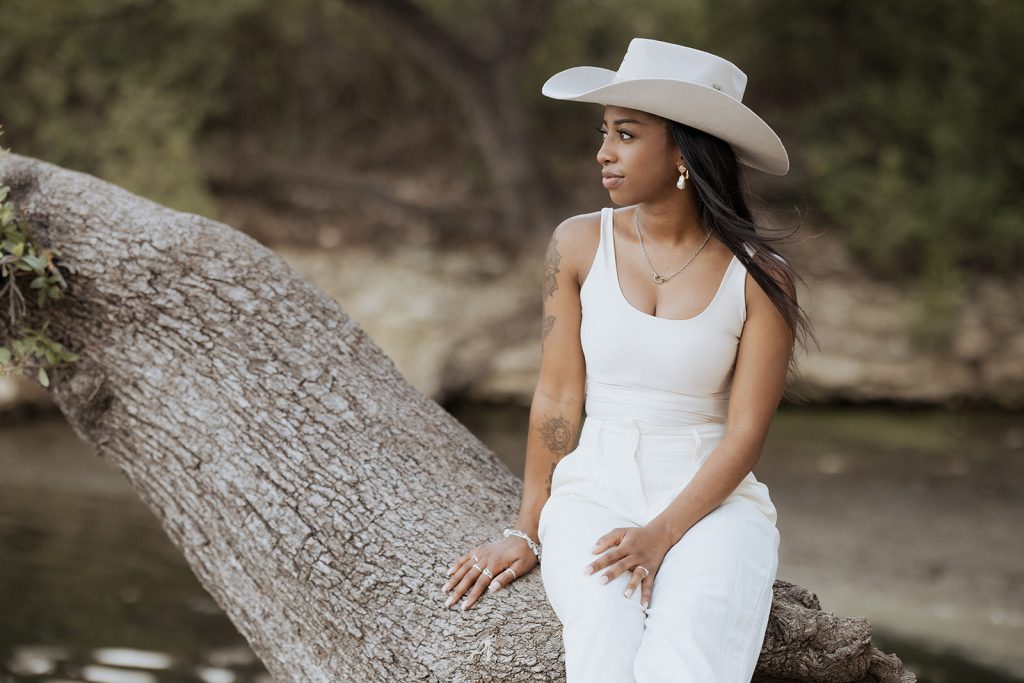 Woman in a light dress standing in front of palm leaves, gently touching her necklace and gazing thoughtfully into the distance in soft natural light.