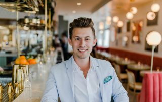 Confident young man in a light blazer smiling while sitting at a stylish bar with a bright and modern interior.