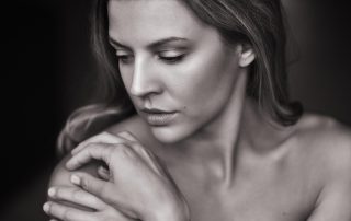 Black and white close-up portrait of a woman with soft lighting, gently holding her shoulder and looking down with a serene expression.