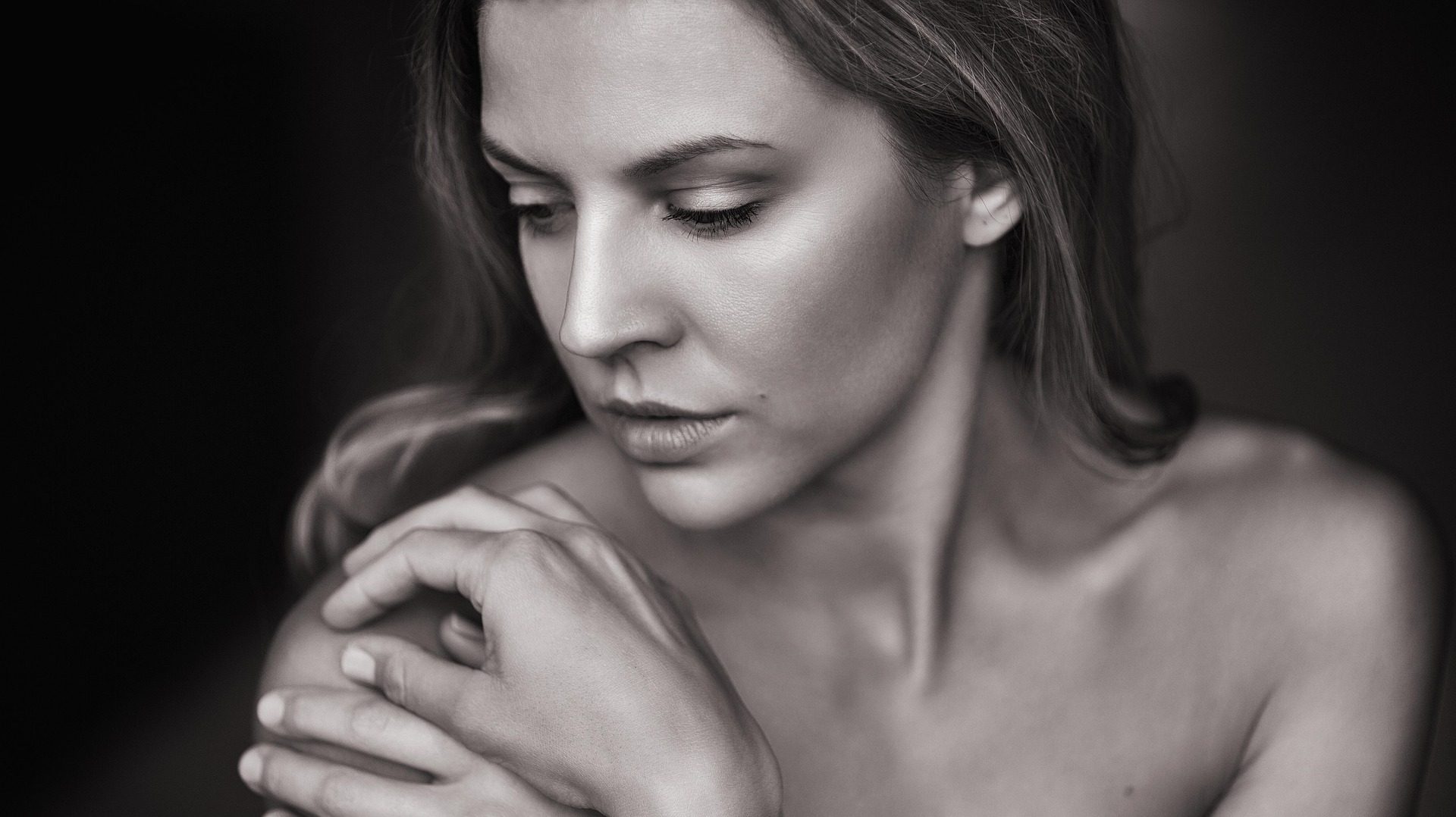 Black and white close-up portrait of a woman with soft lighting, gently holding her shoulder and looking down with a serene expression.