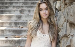 Young woman with long blonde hair wearing a white dress, sitting on stone steps next to a rustic stone wall, softly lit by natural light.