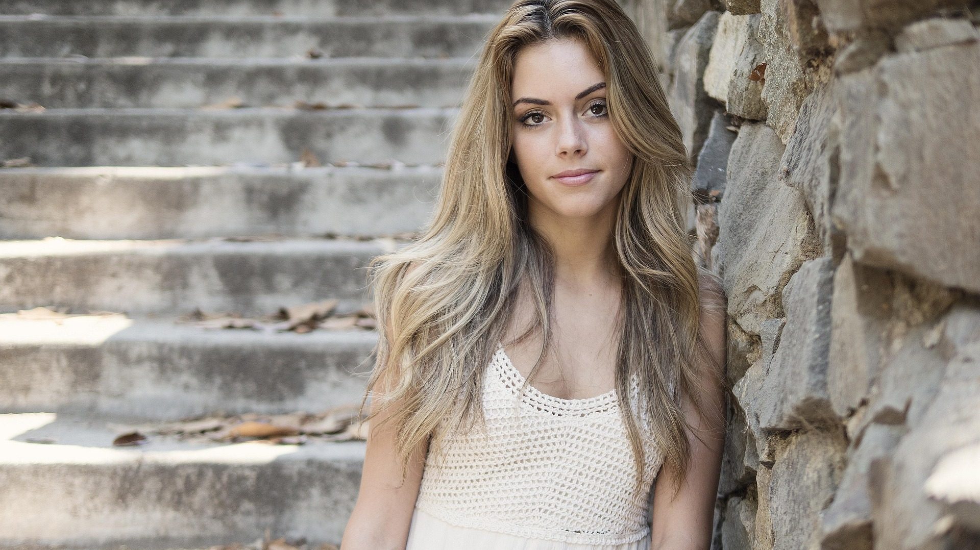 Young woman with long blonde hair wearing a white dress, sitting on stone steps next to a rustic stone wall, softly lit by natural light.