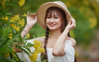 A smiling young woman in a white off-shoulder top and straw hat standing among blooming yellow flowers in a garden.