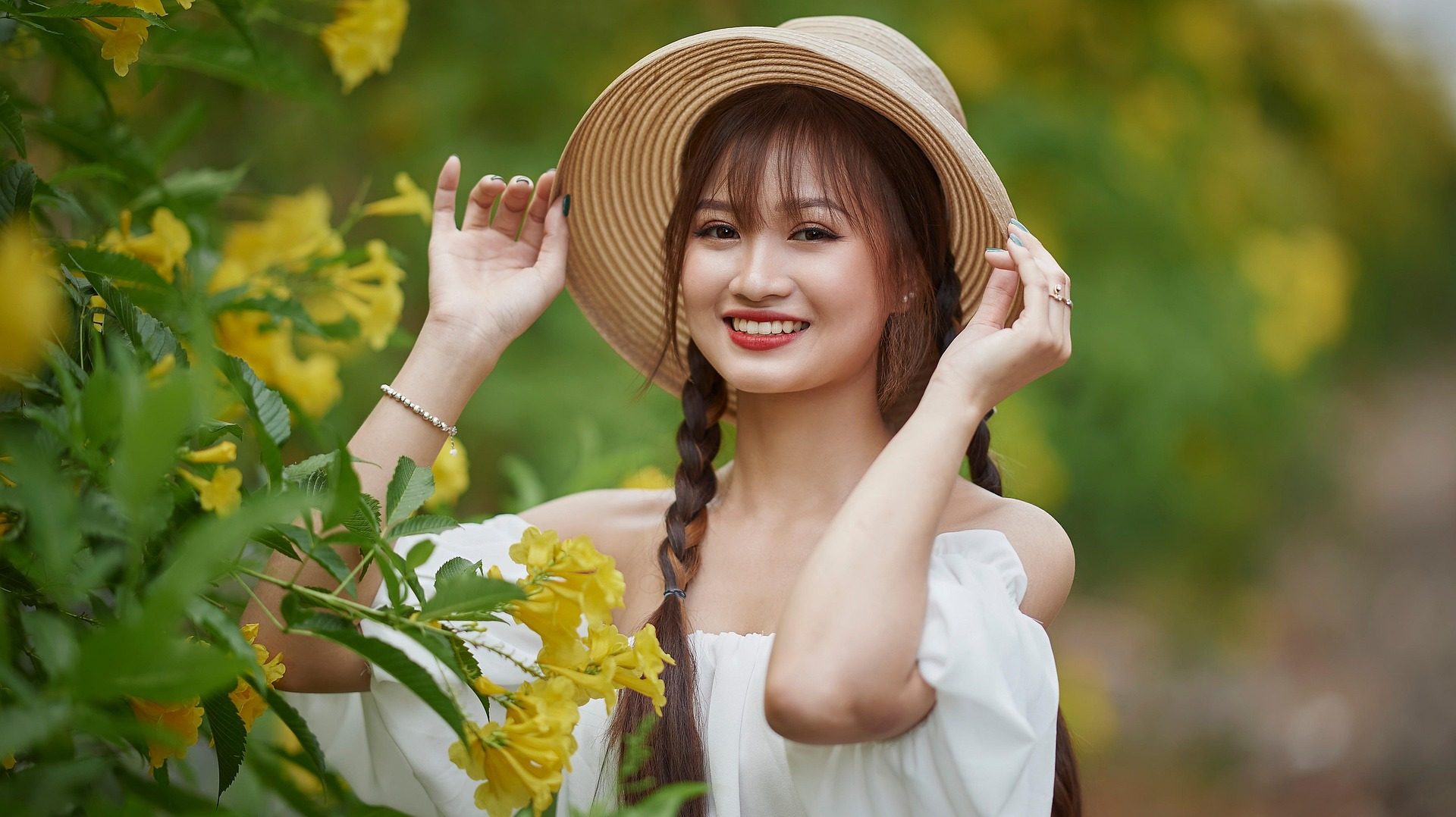 A smiling young woman in a white off-shoulder top and straw hat standing among blooming yellow flowers in a garden.