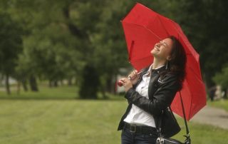A smiling young woman in a leather jacket holding a red umbrella, standing in a green park and looking up with joy.