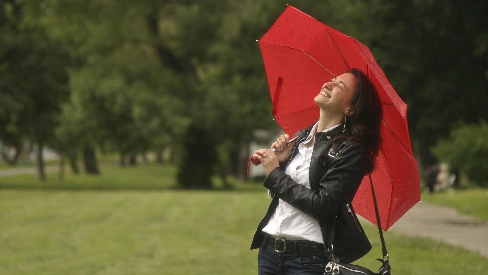 A smiling young woman in a leather jacket holding a red umbrella, standing in a green park and looking up with joy.