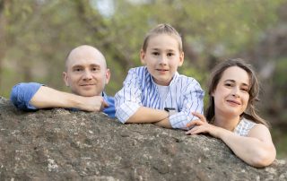 A family of three—father, mother, and young son—leaning over a large rock and smiling outdoors, with soft spring greenery in the background.