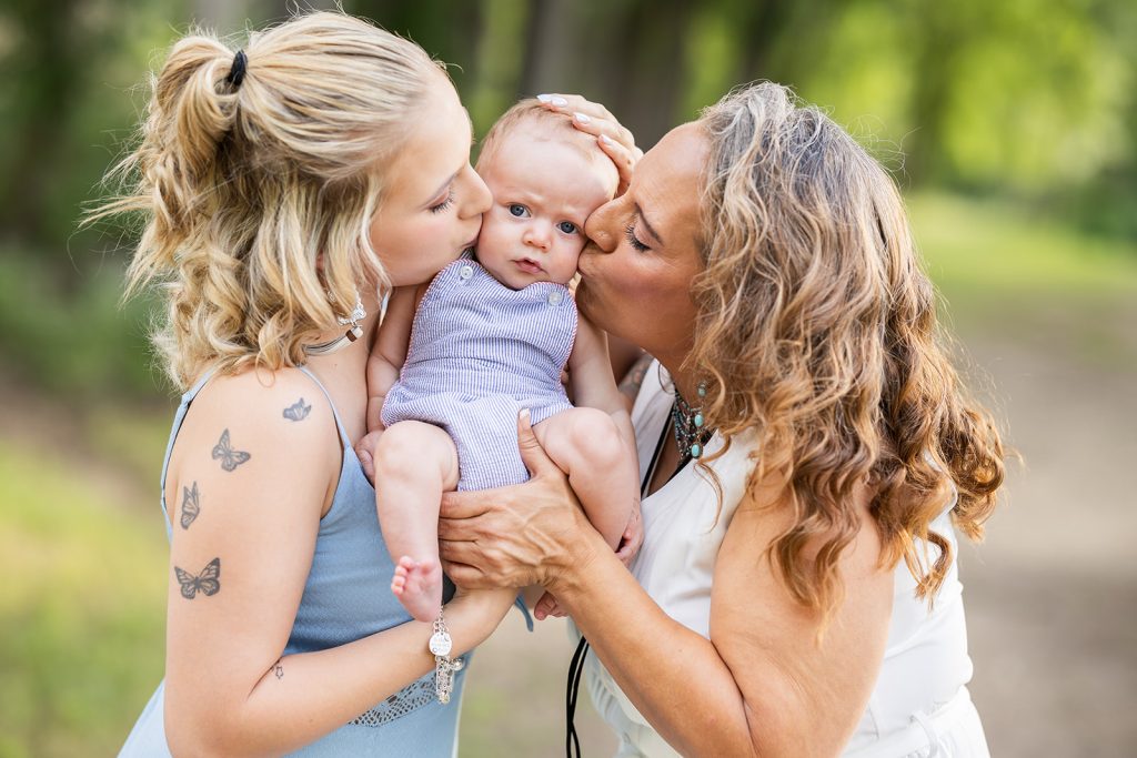 Baby being kissed on both cheeks by mother and grandmother, with a surprised expression.