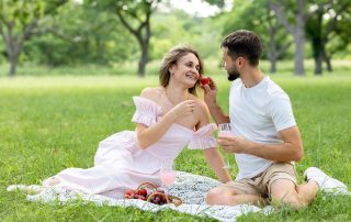 A couple on a picnic in a park. A woman in a pink dress and a man in a white t-shirt are laughing and looking at each other while she offers him a strawberry.