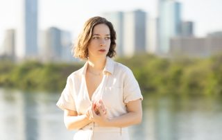 Woman standing by the river with downtown Austin skyline in the background, bathed in soft natural light.