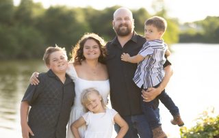 A joyful family of five poses together by the water at sunset, with all members smiling and dressed in light, casual clothing for a warm outdoor session.