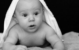 Black and white portrait of a newborn baby lying on a towel with a hooded blanket over their head, looking curiously at the camera.