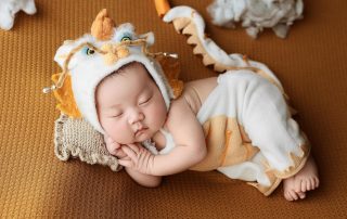 Newborn baby sleeping peacefully on a textured brown blanket, dressed in a whimsical dragon-themed costume with a hat and tail.
