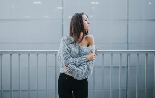 A stylish young woman standing near a metal railing, wearing a loose knit sweater and black pants, looking to the side in a minimalist urban setting.