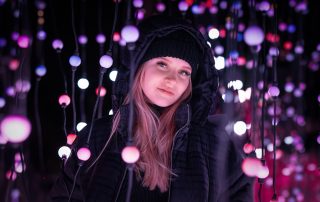 A young woman in a black winter jacket and beanie stands among vibrant hanging lights in shades of purple, pink, and white, creating a dreamy, futuristic atmosphere at night.