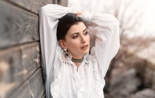 Woman in a white ruffled blouse leaning against a wooden wall, wearing large pearl earrings and looking away thoughtfully.