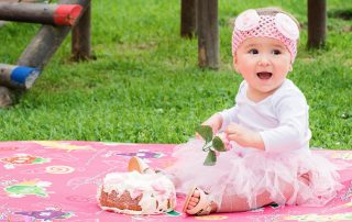 Smiling baby girl in a pink tutu and headband sitting on a colorful mat with a cake during a birthday celebration outdoors.