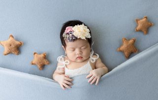 Newborn baby girl sleeping peacefully on a soft blue blanket, tucked in under the fabric with a white knitted top and a colorful flower headband, surrounded by five small plush star pillows.