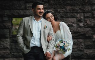 Bride resting her head on groom’s shoulder while holding a bouquet, sitting together by a stone wall in a quiet outdoor setting