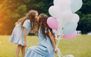 Mother and daughter in matching blue dresses share a kiss while holding pastel balloons in a sunny green field.