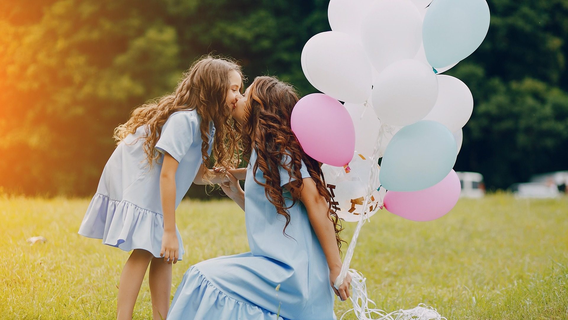 Mother and daughter in matching blue dresses share a kiss while holding pastel balloons in a sunny green field.