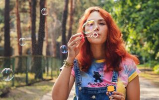 Young woman with red hair blowing soap bubbles in a sunny park while wearing a colorful tie-dye shirt and denim overalls.